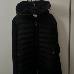Svart Moncler pufferjacka med stickade ärmar - Svart cardigan från Moncler med stickade ärmar och huva. Jackan har dragkedja framtill, två fickor med tryckknappar och Moncler-logga på ärmen. Beställde den från Monclers hemsida så har fysiskt kvitto och digitalt kvitto. Använt få gånger, säljer för den inte passar mig.