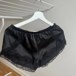Svarta silkesliknande shorts med spets - Snygga svarta shorts i silkesliknande material med bred spetskant längs benen och dekorativa spetsdetaljer framtill. De har elastisk midja och en loose passform, perfekta för chill eller som pyjamasshorts. Riktigt mjuka och sköna!