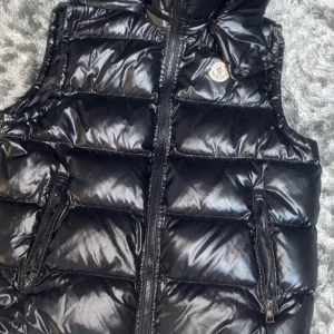 Moncler väst - Säljer en svart, glansig dunväst från Moncler med klassisk logga på bröstet. Västen har två fickor med dragkedja och quiltad design. Perfekt att slänga över en hoodie för en cool look. Insidan har en unik Moncler-serieetikett.