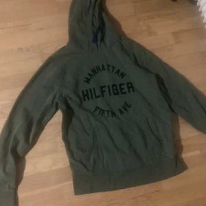 Grön hoodie från Hilfiger Manhattan - Säljer en grön hoodie från Hilfiger med trycket 'Manhattan Fifth Ave' på bröstet. Tröjan har huva med snörning och en stor magficka. Perfekt för chill dagar och streetwear-stil. Obs man får båda på samma köp. Tröjan är M