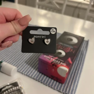 Montini örhängen hjärtformade silver - Snygga örhängen från Montini i hjärtformad design med silverfärgad kant och vit insida. Varje hjärta har en liten glittrande sten i mitten som ger extra bling. Perfekt accessoar för att lyfta din look med en söt detalj. Aldrig provade.