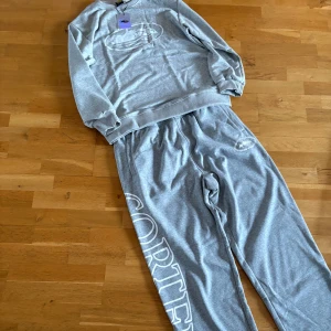 Grå mjukisbyxor och tröja från Corteiz - Grå mjukisbyxor från Corteiz med stor vit logotyp längs benet och tryck vid fickan. Byxorna har elastisk midja och är gjorda i mjukt bomullsmaterial för maximal komfort. Perfekt för chill dagar och streetwear-stil.