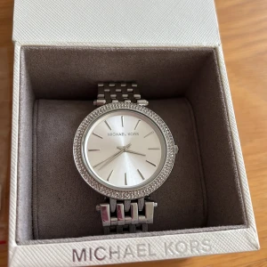 Silverfärgad klocka från Michael Kors - Stilren klocka från Michael Kors i silverfärg.  Jag skickar med en länk till klockans armband så att man kan justera armbandets storlek runt handleden. 