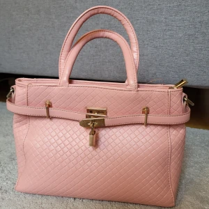 Rosa handväska med guldiga detaljer - Säljer en snygg rosa handväska med guldiga metalldetaljer. Väskan har två handtag, dekorativt lås framtill och ett stilrent rutmönster. Perfekt för dig som vill ha en chic och trendig accessoar till din outfit. Axelrem tillkommer. Väskan är ej använd mycket alls, men litet slitage finns på ett av handtagen efter förvaring.