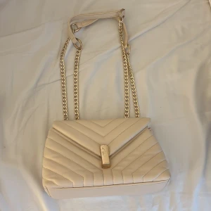 Beige quiltad axelväska med kedja - Snygg beige axelväska med quiltad design och guldfärgad kedjerem. Väskan har ett elegant spänne framtill och är tillverkad i skinnimitation. Perfekt storlek för det viktigaste och ger en lyxig känsla till din outfit.