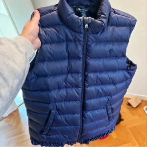 Ralph Lauren väst - Snygg marinblå dunväst från Polo Ralph Lauren som passar storlek xs-s. Västen har quiltad design, hög krage, dragkedja framtill och två fickor. Klassisk broderad logga på bröstet. Perfekt för lager-på-lager under höst och vinter.
