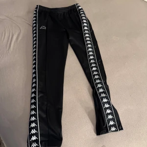 Svarta Kappa träningsbyxor med logga - Svarta träningsbyxor från Kappa med klassisk vit logga längs båda sidorna och vita detaljer. Byxorna har elastisk midja och tryckknappar längs benen för extra stilpoäng. Perfekta för dig som gillar sportig och retro vibe.