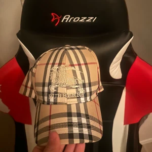 Burberry rutig keps i beige och svart - Snygg keps från Burberry med klassiskt rutigt mönster i beige, svart och rött. Framtill finns ett broderat Burberry-emblem med riddare och logga. Kepsen är tillverkad i bomull och har justerbar passform. Perfekt för dig som vill sticka ut med en ikonisk accessoar.