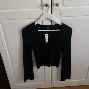 Svart ribbad kofta från Gina Tricot - Svart kofta från Lindex  med ribbad struktur och v-ringad hals. Koftan har långa ärmar och knappar framtill. Perfekt att styla med jeans till ett linne. Den är helt oanvänd och helt ny. Den köptes för 499kr i mitten av sommaren.