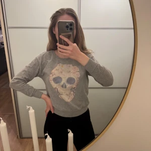 Grå sweatshirt med döskalle från H&M Divided - Grå sweatshirt från H&M Divided i storlek XS. Tröjan har ett stort tryck av en döskalle med blommor framtill, rund halsringning och ribbade muddar vid ärmslut och nederkant. Perfekt för dig som gillar edgy och cool stil.  * Fint skick - lite nopprig se bild men man ser bara om man tittar noga * Skickar samma dag eller dagen efter  *  Rökfritt hem