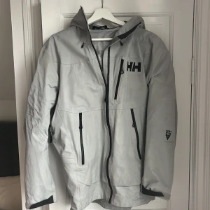 Ljusgrå vindjacka från Helly Hansen - Säljer en ljusgrå vindjacka från Helly Hansen med huva och svarta detaljer. Jackan har flera dragkedjeförsedda fickor, justerbara ärmslut och HH-logga på bröstet och huvan. Perfekt för skidåkning 