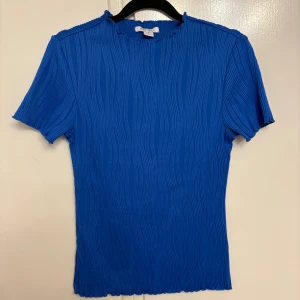 Blå ribbad topp från Amisu - Säljer en blå ribbad topp från Amisu i storlek S. Toppen har korta ärmar, rund hals med vågig kant och ett snyggt strukturerat tyg med vertikala ränder. Perfekt för dig som gillar färg och vill ha en enkel men cool look.