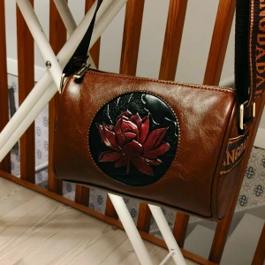 Axelväska med svart och rött mönster - Helt ny axelväska i skinnimitation med en stor, präglad röd blomma på framsidan. Väskan har justerbar axelrem med text, dragkedja upptill och rymligt fack. Perfekt för dig som vill sticka ut med en unik accessoar.