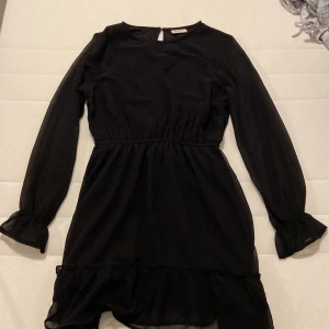 Svart volangklänning från Kappahl - Svart klänning från Kappahl med långa ärmar och volangdetalj nertill. Klänningen har markerad midja och lätt transparenta ärmar med liten volang vid ärmslut. Enkel och stilren design, perfekt för dig som gillar klassiska plagg.