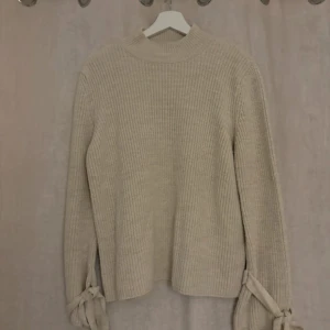 Beige stickad tröja med knyt från Gina Tricot - Superfin beige stickad tröja från Gina Tricot med ribbad struktur och snygga knytband vid ärmsluten. Tröjan har en högre rund halsringning och långa ärmar, perfekt för lager på lager under kyliga dagar.