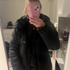 Svart dunjacka från Boomerang - Säljer en svart, klassisk dunjacka från Boomerang med huva och snygga detaljer som knappar och dragkedja. Jackan är fluffig och har en skön passform, perfekt för kalla dagar. Modellen är långärmad och har en stilren look som funkar till de flesta outfits. storlek M, de saknas en knapp men är lätt att sy dit en ny