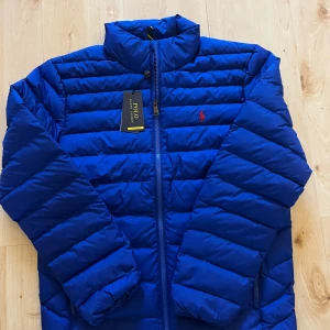 Blå pufferjacka från Polo Ralph Lauren - Snygg blå pufferjacka från Polo Ralph Lauren med röd broderad logga på bröstet. Jackan har hög krage, dragkedja framtill och två sidofickor med dragkedja. Perfekt för kyliga dagar och riktigt skön att bära. Färgen är royal blue. Hör av er vid frågor eller funderingar! 