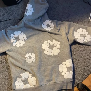 Grå hoodie med vita blommor Denim tears - Säljer en grå hoodie med stora vita blommiga tryck över hela tröjan. Tröjan har huva och en klassisk magficka framtill. Perfekt för dig som vill sticka ut med en cool och unik stil. Mjuk och skön att ha på sig.