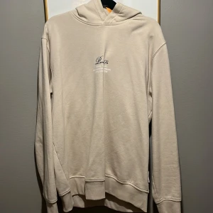 Beige hoodie Paris Fr tryck - Snygg beige hoodie med tryck 'Paris, Fr' och text om olika stadsdelar i Paris både fram och bak. Mjuk bomullskänsla, långärmad och med huva. Perfekt för dig som gillar stilrena plagg med coolt print.