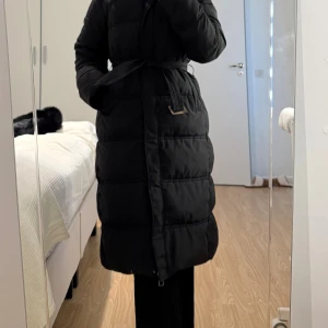 Svart lång pufferjacka med bälte - Säljer en svart lång pufferjacka med huva och avtagbar fuskpäls. Jackan har bälte i midjan för snygg passform och stängs med dragkedja. Perfekt för kalla dagar och riktigt mysig att bära. Klassisk look som funkar till allt.