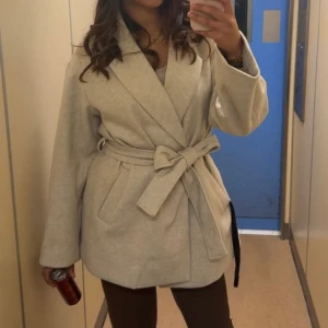 Beige omlottkappa med bälte - Säljer en stilren beige kappa i omlottmodell med bälte i midjan. Jackan har bred krage och långa ärmar, perfekt för dig som vill ha en klassisk och trendig look. Passar snyggt till både jeans och klänning.