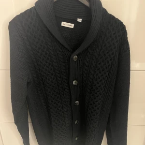 Svart stickad kofta från Jack & Jones - Säljer en svart stickad kofta från Jack & Jones med snyggt kabelstickat mönster framtill och bred krage. Koftan har knappar hela vägen och långärmade ärmar. Perfekt för dig som gillar klassisk stil med modern touch. Aldrig använd då den var för stor för mig, är i helt nytt skick