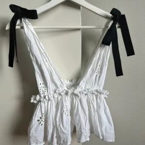 Vit blus med svarta rosetter - Supersöt vit blus med broderade hålmönster och volangkant. Axelbanden knyts med stora svarta rosetter för en extra gullig touch. Blusen har djup v-ringning och är ärmlös, perfekt för varma dagar och en trendig look.