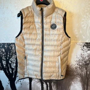 Beige dunväst från Pelle P, M - Snygg beige dunväst från Pelle P med quiltad design och hög krage. Västen har svart kant vid ärmhålen, två fickor med dragkedja och logotyp på bröstet. Perfekt för lager-på-lager och har en stilren look med diskret glansigt material.