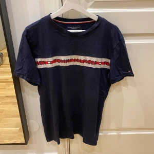 Tommy Hilfiger t-shirt - Snygg mörkblå t-shirt från Tommy Hilfiger med klassisk logga i rött och vitt över bröstet. T-shirten har rund hals och korta ärmar. Materialet är mjuk bomull som känns skönt mot huden. Perfekt för dig som gillar stilrena och ikoniska plagg.