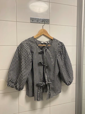 Rutig blus med knyt från Kappahl - En svartvit rutig blus från Kappahl i storlek XS. Blusen har puffiga långärmar och flera snygga knyt framtill som ger en cool touch. Tillverkad i mjuk bomull, perfekt för dig som gillar statement-plagg med retrovibe. Använts enbart en gång och utan defekter. 
