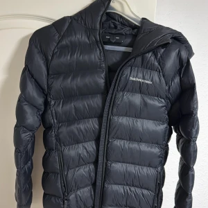 Svart dunjacka Peak Performance  - Säljer en svart dunjacka från Peak Performance i storlek M. Jackan har huva, dragkedja framtill och två sidofickor med dragkedja. Klassisk pufferstil med diskret logga på bröstet och ärmen. 