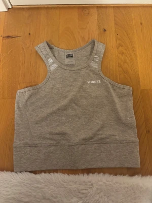 Biege linnetopp från Stronger - Säljer en biege linnetopp från Stronger med vita detaljer vid axlarna och logga på bröstet. Toppen är ärmlös och har en sportig, croppad passform som passar perfekt till träning. Materialet är mjukt och stretchigt polyester för maximal komfort.