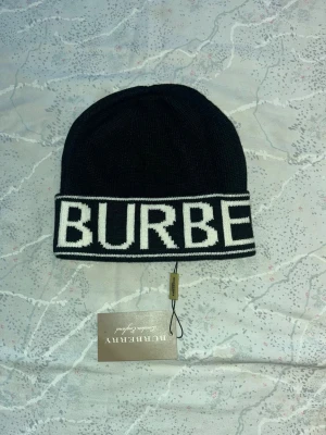 Svart Burberry mössa med vit logga - Snygg svart mössa från Burberry med bred vit kant där BURBERRY är stickat i stora bokstäver. Klassisk och stilren design som passar perfekt till kalla dagar. Mössan är stickad och har en mjuk känsla, med märkets taggar kvar.