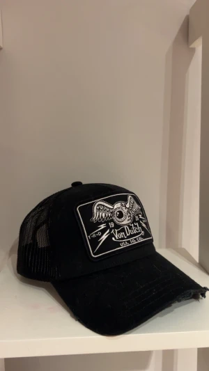 Svart Von Dutch truckerkeps - Svart truckerkeps från Von Dutch med vit broderad patch framtill som har ett coolt öga med vingar och logga. Kepsen har böjd skärm och mesh på baksidan för extra ventilation. Perfekt för dig som gillar streetstyle och vill sticka ut.