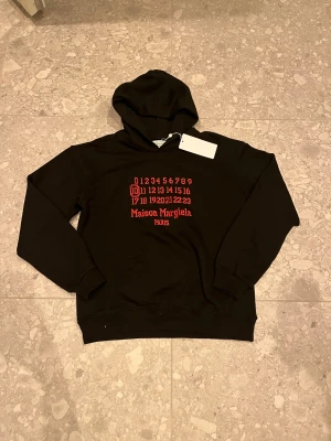 Maison Margiela svart hoodie med röd print - Svart hoodie från Maison Margiela med ikonisk röd print framtill och huva. Snyggt tryck med siffror och logga i rött. Mjuk bomullskvalitet och klassisk passform för denna stil . Perfekt för dig som gillar streetwear och designer. Hör mer än gärna av dig vid mista fråga😁