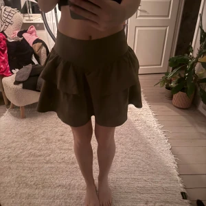 Khaki volangkjol med hög midja - Supersnygg khaki kjol med volanger och hög midja. Kjolen är kort och har en skön, lite loose passform. Perfekt för dig som gillar en feminin och trendig look. Materialet känns mjukt och följsamt mot kroppen. Märke är oklart