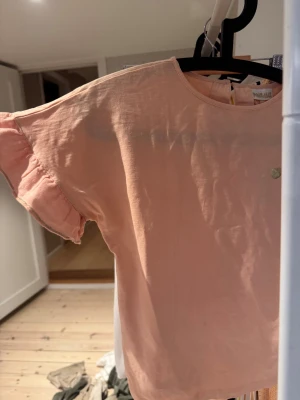 Puderrosa blus med volangärm Pompdelux - Supersöt puderrosa blus från Pompdelux i mjuk bomull. Blusen har rund hals och korta ärmar med volangkant och diskret glittrig söm. Perfekt för dig som gillar romantiska detaljer och en chill vibe. Färgen är ljus och passar till allt. Prislappen är kvar, helt ny. Säljer för 85kr. Passar från 10 - 12 åring. Strl 134/140. Kontakta mig för bättre bilder