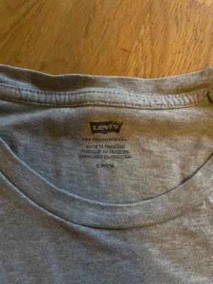 Grå Levi's t-shirt med röd logga - Snygg grå t-shirt från Levi's med klassisk röd logga på bröstet och liten röd etikett i sidan. T-shirten är i mjuk bomull och har rund hals och korta ärmar. Perfekt för en avslappnad och stilren look.