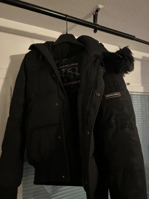 Svart pufferjacka Kings Will Dream - Svart pufferjacka från Kings Will Dream med huva och snygg pälsdetalj. Jackan har dragkedja framtill, diskret camo-mönster på ärmarna och ribbade muddar. Perfekt för kalla dagar och har en cool streetstil. Insidan är fodrad med mjukt material för extra komfort.