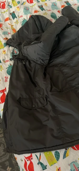 Mörk grå pufferjacka med huva - Säljer en mörk grå pufferjacka med huva och flera fickor med både dragkedja och knappstängning. Jackan har långa ärmar och justerbar nederkant med knappar. Perfekt för kalla dagar och har en clean, stilren look.