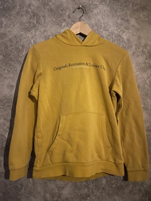 Gul hoodie från Jack & Jones - Gul hoodie från Jack & Jones med trycket 'Originals Recreation & Leisure Club' på bröstet. Tröjan har huva, magficka och är tillverkad i mjuk bomull. Perfekt för dig som gillar en avslappnad och stilren look.