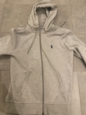 Grå hoodie från Polo Ralph Lauren - Snygg ljusgrå hoodie från Polo Ralph Lauren med dragkedja och huva. Tröjan har två fickor framtill, snörning vid huvan och den klassiska Polo-loggan broderad på bröstet. Perfekt för chill dagar och enkel att matcha med jeans.