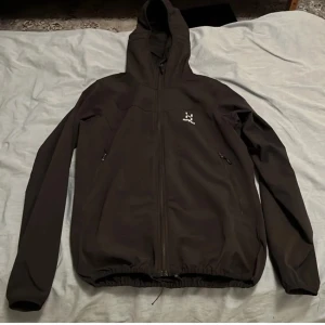 Svart vindjacka från Haglöfs - Svart vindjacka från Haglöfs med huva och hel dragkedja framtill. Jackan har två sidofickor med dragkedja och justerbar nederkant med snörning. Perfekt för blåsiga dagar och outdoor-aktiviteter. Diskret Haglöfs-logga på bröstet.