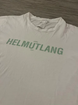 Vit Helmut Lang t-shirt med tryck - Snygg vit t-shirt från Helmut Lang med grönt logotryck på bröstet. Klassisk rund hals och korta ärmar. Tillverkad i 100% bomull för skön och lätt känsla. Perfekt för dig som gillar minimalistisk design och streetwear-vibe. Något missfärgad under ärmarna. Hör gärna av er vid prisförslag eller andra frågor!