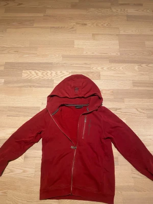 Röd hoodiejacka från Sail Racing - Röd hoodiejacka från Sail Racing med dragkedja och broderad logga på huvan. Jackan har en bröstficka med dragkedja och mjukt fleecefoder på insidan. Snygga svarta och vita detaljer vid kragen. Perfekt för chilliga dagar. Pris kan diskuteras!