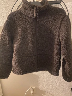 Brun teddyjacka från Gina Tricot - Mysig brun teddyjacka från Gina Tricot i storlek 158/164. Jackan har hög krage, hel dragkedja framtill och långärmade ärmar. Materialet är fluffigt och ger en cool, avslappnad vibe. Perfekt för dig som gillar att hålla dig varm och ändå se chill ut.