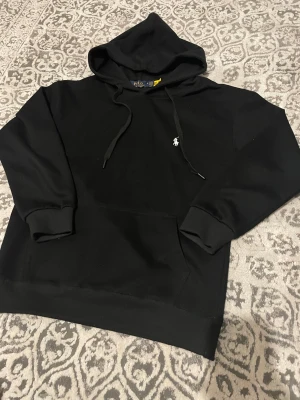 Svart hoodie från Polo Ralph Lauren - Svart hoodie från Polo Ralph Lauren med klassisk vit broderad logga på bröstet. Tröjan har huva med snörning, stor magficka och ribbade muddar vid ärmar och nederkant. Tillverkad i 100% bomull för skön och mjuk känsla. Perfekt för chill dagar.
