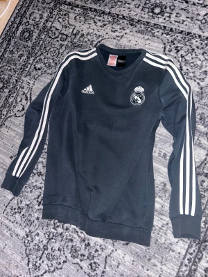 Real Madrid tröja - Svart långärmad tröja från Adidas med Real Madrid på bröstet och klassiska vita ränder längs ärmarna. Crewneck och mjukt material som är skönt mot huden. Perfekt för dig som gillar fotboll och vill ha en sportig stil.