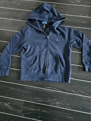 Mörkblå hoodie från Polo Ralph Lauren - Mörkblå hoodie från Polo Ralph Lauren med dragkedja framtill och klassisk huva. Tröjan har en röd broderad logga på bröstet och två fickor framtill. Perfekt för en chill och stilren look.