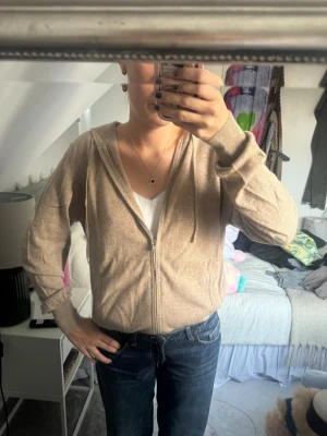 Beige/brun hoodie i kashmir från Soft Goat - Mjuk, beige/brun hoodie från Soft Goat i storlek L, tillverkad i kashmir. Tröjan har dragkedja framtill, huva med snörning och långa ärmar. 